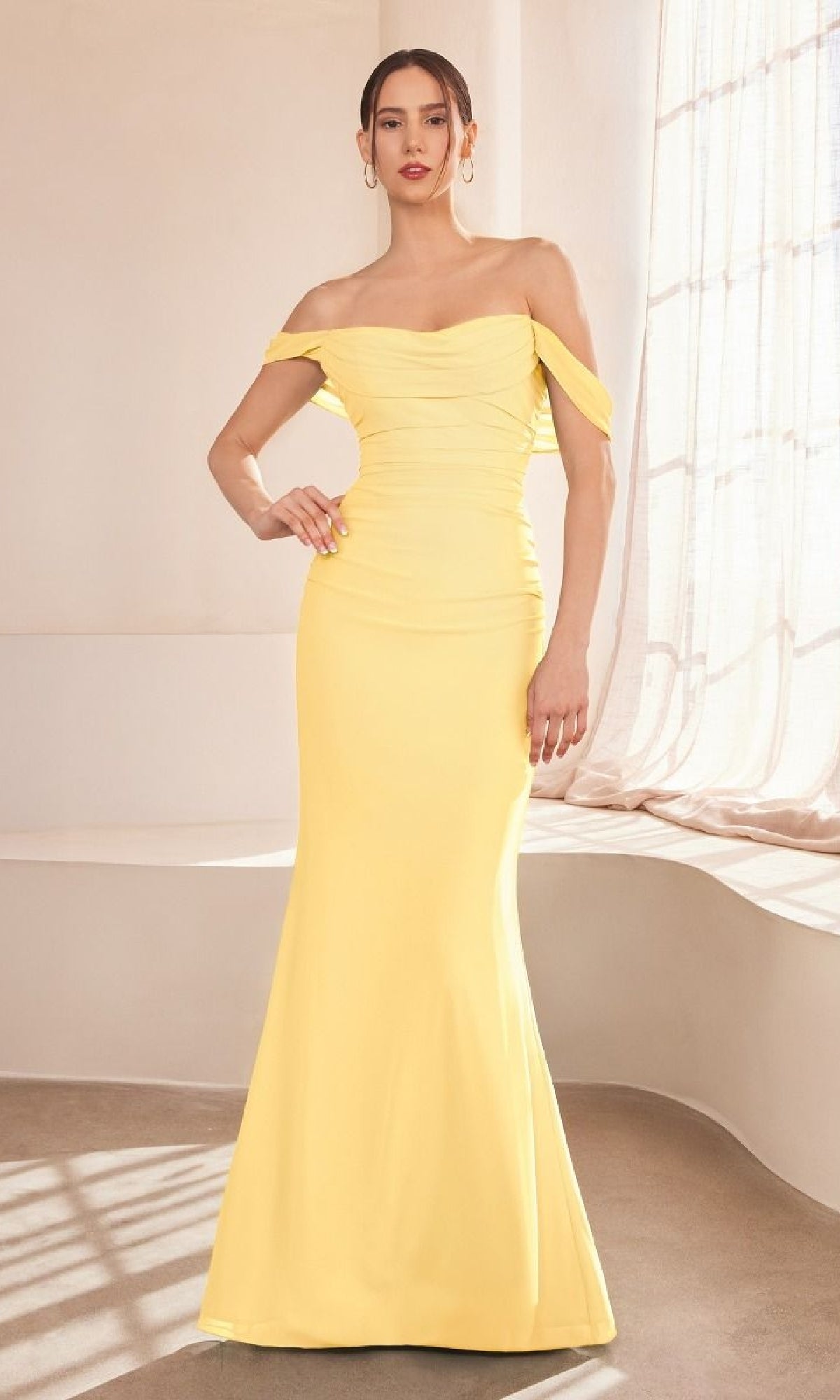 A woman in the affordable Amoris T705 Off-Shoulder Classic Prom Gown stands by a sunlit window, her yellow floor-length dress glowing softly through sheer curtains.